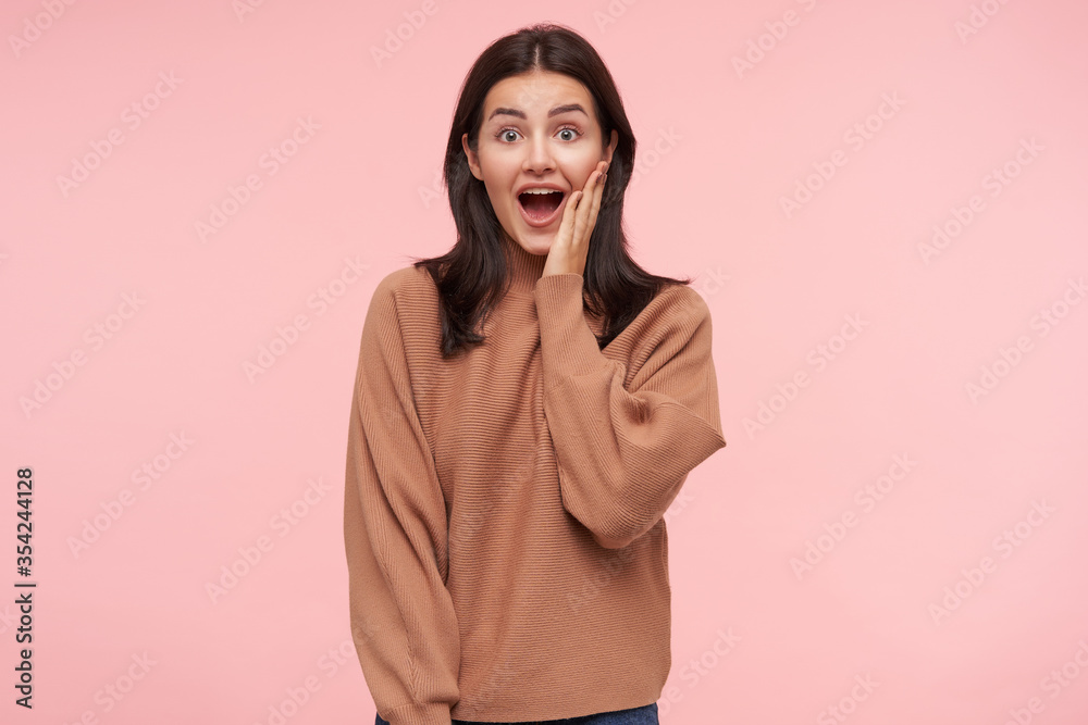 Wall mural Bemused young lovely brunette female with casual hairstyle keeping her mouth wide opened while looking dazedly at camera, raising hand to her face while posing over pink background