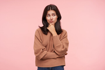 Bewildered young attractive brown haired lady dressed in knitted roll-neck sweater touching her chin with raised hand and looking thoughtfully aside while posing over pink background
