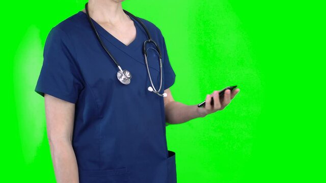 4K female nurse doctor on green screen isolated with chroma key. Woman looks at her smartphone, puts it in her pocket and stands with arms crossed
