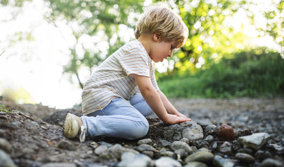 Side view of small boy playing with rocks and mud in nature. - obrazy, fototapety, plakaty