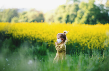 Side view of small toddler girl with face mask standing in nature in rapeseed field.