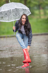 Woman laughing at water leaking to her rain boot, taking it off, while she keeps protecting herself againt the rain by an umbrella