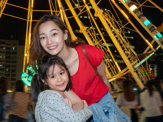 happy asia mother and daughter have fun in amusement carnival park with farris wheel and carousel...