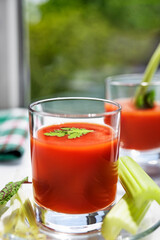 Two glasses of tomato juice with parsley and celery decorations, stand near the window, morning sunlight shines, shallow depth of field, selective focus. Natural drinks concept.