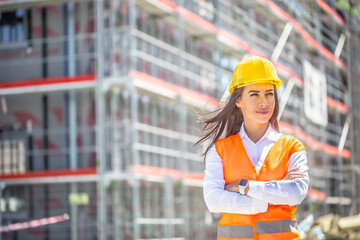 Female site supervisor wearing safety vest and helmet stands confidently in front of the construction covered in scaffolding