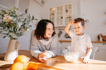 Mom with her son is cooking on the background of a bright kitchen. The child helps parents knead the dough for baking in conditions of self-isolation.The boy lifted a whisk.