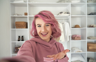 Happy hipster gen z teen girl fashion social media channel blogger stylist with pink hair wearing hoodie looking at camera recording vlog video tutorial in front of clothes wardrobe, face headshot.