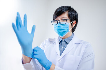 Male Asian doctor or Physician man in white uniform and blue surgical mask putting disposable latex gloves on hand while working in hospital clinic. Coronavirus (COVID-19) infection prevention.