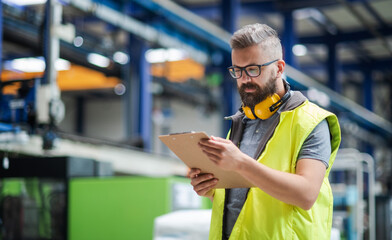 Technician or engineer with protective headphones standing in industrial factory.