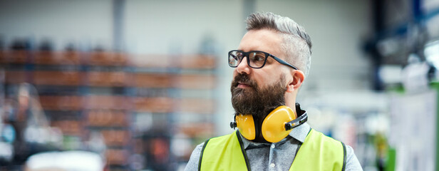 Technician or engineer with protective headphones standing in industrial factory.