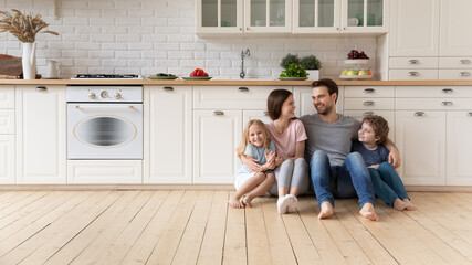 Happy young family with little children sit on warm wooden floor in new modern design kitchen,...