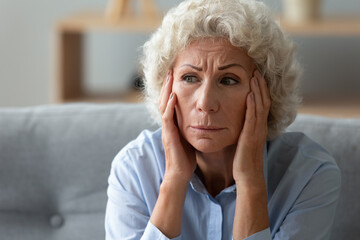 Close up worried elderly 70s woman seated on sofa touch head with hands deep in sad thoughts,...