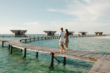 Back view couple walking together on wooden jetty near wter bungalows. Couple in white on a...
