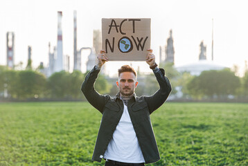 Young activist with placard standing outdoors by oil refinery, protesting.