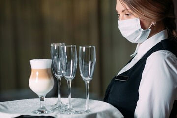 A female Waiter of European appearance in a medical mask serves Latte coffee.