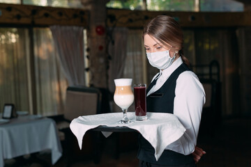 A female Waiter of European appearance in a medical mask serves Latte coffee.