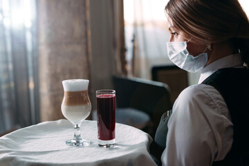 A female Waiter of European appearance in a medical mask serves Latte coffee.