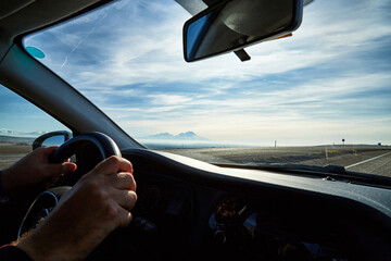 Fototapeta premium Windscreen view of nature, road, sky and hand of man on the steering wheel in a spring, summer or autumn day. The concept of a single trip