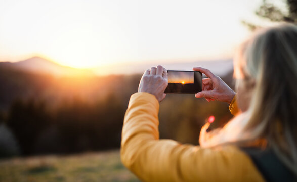 Senior woman hiker standing outdoors in nature at sunset, taking photograph.