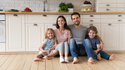 Portrait of young happy family with small children relax on wooden floor in new design kitchen, smiling Caucasian parents rest enjoy leisure weekend with little kids in rent renovated home or studio