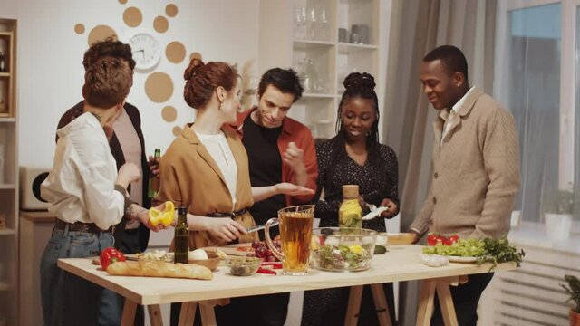 Company of young diverse friends smiling and joking while getting ready for home dinner together: cheerful women cooking food while their boyfriends having beer and stealing some vegetables