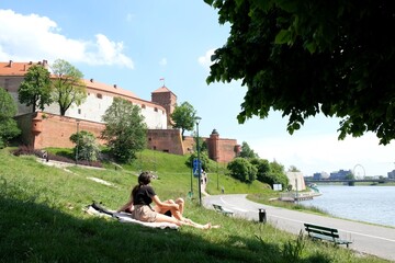 Fototapeta The girls are sitting on a blanket on the grass by the Vistula River next to the Wawel Royal Castle. People are walking along the river. obraz