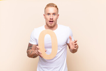 young blonde man surprised, shocked, amazed, holding a number 0.