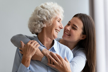 Loving cheerful grown up daughter hugs elderly mother from behind, multi generational relatives beautiful women enjoy time together laughing having fun, love care understanding concept, close up image