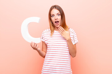 young pretty red head woman surprised, shocked, amazed, holding the letter C of the alphabet to...