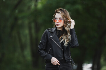 Young beautiful girl in a leather jacket and fashionable sunglasses is walking alone outdoors in summer day