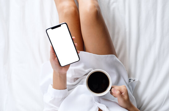 Top view mockup image of a woman holding white mobile phone with blank desktop screen while drinking coffee on a cozy white bed at home in the morning
