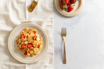 Tiny pancake cereal with strawberries on a plate on white concrete background. Trendy food. Mini cereal pancakes. Trendy breakfast during quarantine. Top view, copy space. Restaurant menu, recipe