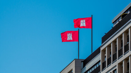 two red flags of hanseatic city of hamburg with coat of arms are waving in the wind on top of an office building. horizontal photography