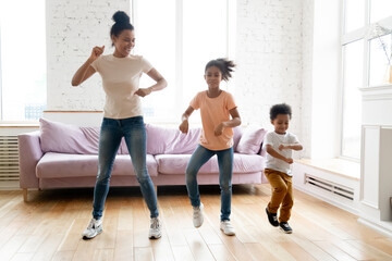 Active mother teaches little children son and daughter to dance, lively African ethnicity family fooling around moving enjoy weekend having fun together in modern living room at home, full length view