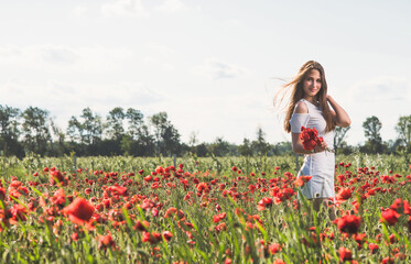 Obraz na płótnie Canvas A dark, stylish, slender girl poses against the background of a poppy field with her hair flowing in the wind in the warm rays of the summer sun