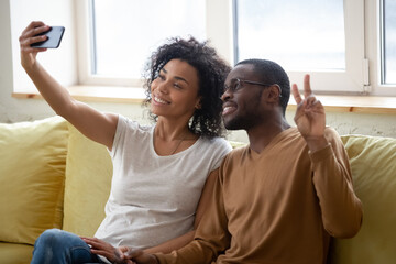 African couple in love sitting on couch using smart phone taking self-portrait having fun making selfie photo for social network memory capture moment. Modern wireless technologies and devices concept