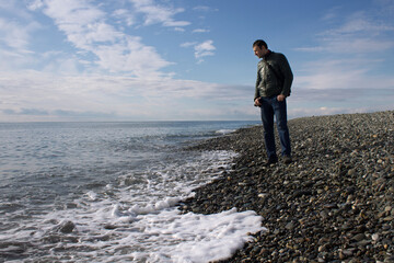 A man dressed in blue jeans and a jacket stands on a rocky beach and looks at the edge of the surf, thinking about something. © Ekaterina