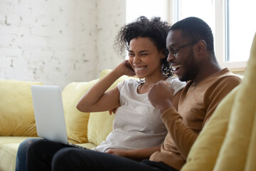 Bank loan application for African family is approved concept. American spouses excited wife and husband sit on couch read received notice on computer feel happy, celebrate success, on-line lottery win