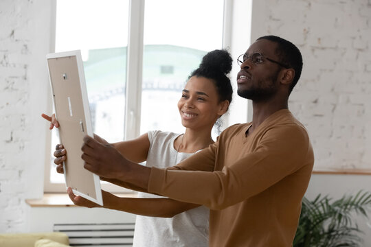 African ethnicity millennial couple holding frame admires painting or photo at relocation day at new first home. Concept of remodeling renovation, new furniture store, bank loan, tenancy rented flat