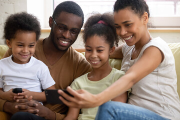 Full african family sit on sofa looking at smartphone screen having fun watch funny videos cartoons on-line. Couple little kids take selfie photo, make videocall chatting with relatives by app concept