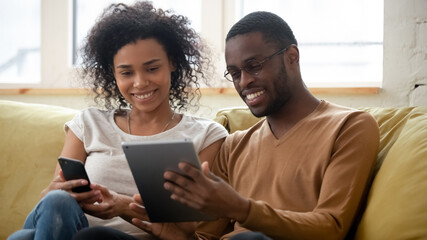 African couple sit on sofa having fun using gadgets. Wife spend time with cell husband holding tablet device show to beloved woman cool app, new video. Modern tech everyday usage in our life concept