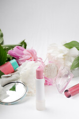 Vertical image.Opened pink lipstick tube, eyeshadow, mirror, liquid lipstick, tube of facial cream and fresh spring peonies on the white surface