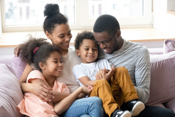 African ethnicity parents pre-teen daughter little son having fun sitting on couch tickling each...