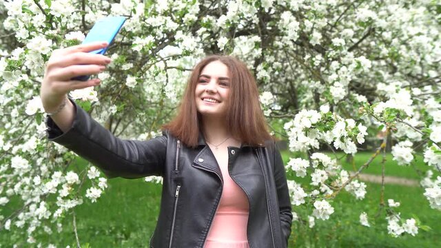 A girl in a dress is photographed in the garden. Photo session in a blooming garden. A beautiful girl is photographed on the phone.A girl in a dress takes a selfie. Lifestyle. 4K.