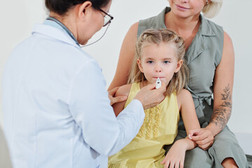Pediatrician putting electronic thermometer in mouth of little girl to check her temperature