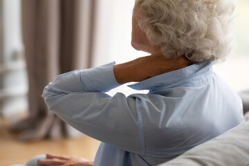 Tired elderly 70s woman sit on sofa massaging stiff sore neck tensed muscles reduces painful...