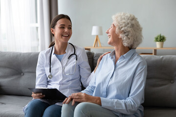 Friendly young nurse in white coat holding clipboard laughing with elderly female patient during...