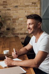 Side view of smiling man holding smartphone near paperwork and books on kitchen table