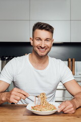 Selective focus of handsome man smiling at camera while eating noodles in kitchen