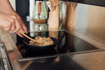 Cropped view of man holding wooden spatula while preparing noodles in frying pan in kitchen